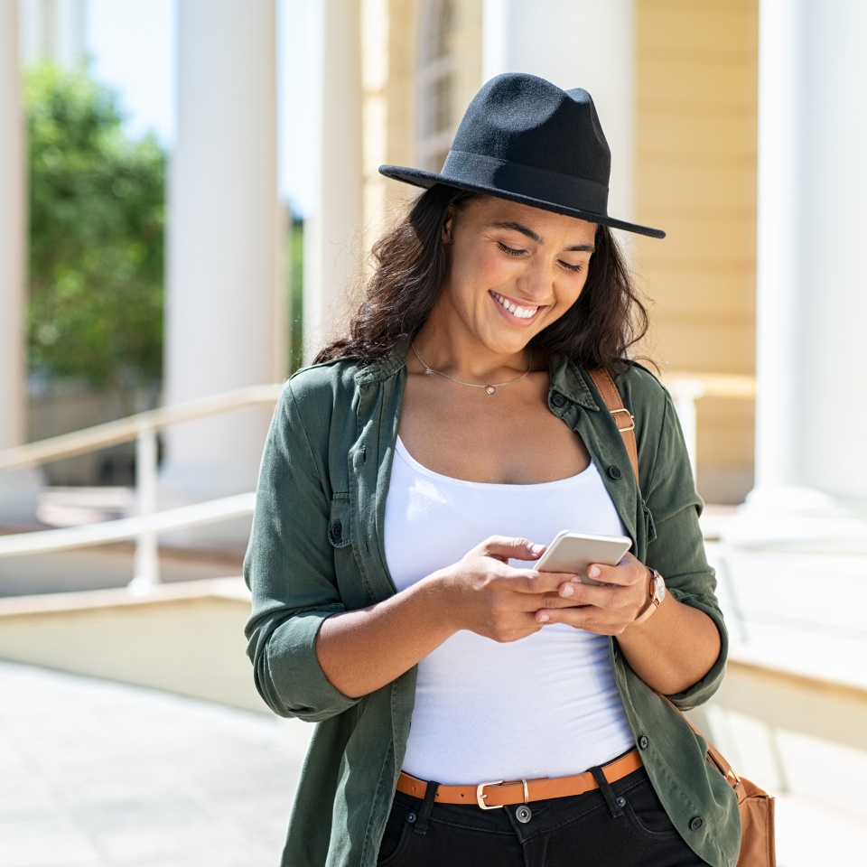 Cheerful young woman wearing black hat in city street typing a message. Hispanic trendy girl browsing internet on phone outdoor. Happy stylish girl using smartphone while travelling the city.