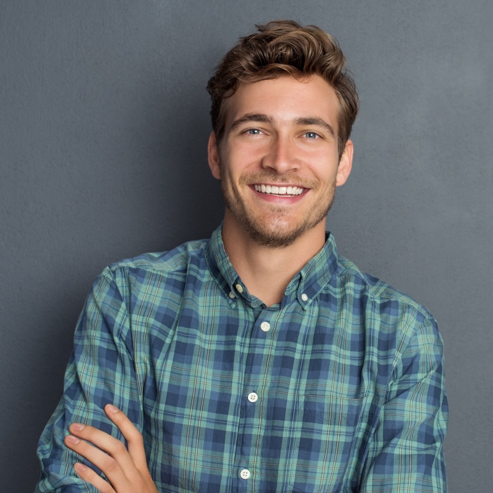 Young handsome man leaning against grey wall with arms crossed. Cheerful man laughing and looking at camera with a big grin. Portrait of a happy young man standing over grey background.