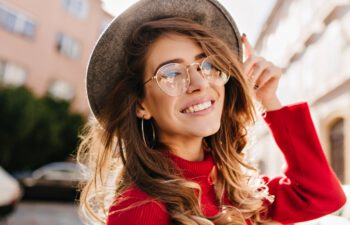 Close-up portrait of cheerful white woman in glasses touching her hat on blur background. Photo of fashionable girl with beautiful brown hair smiling to camera.