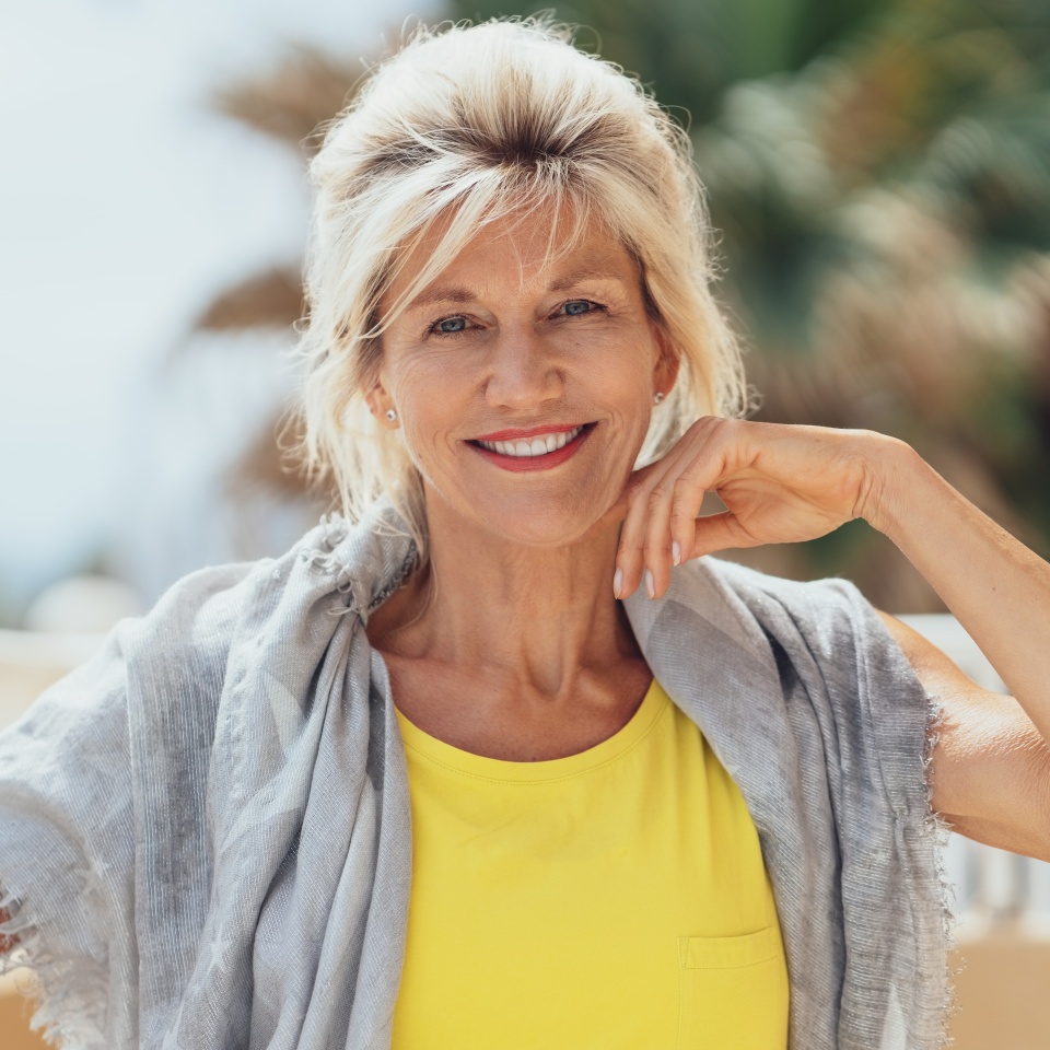 Stylish blond woman sitting on a balcony with a shawl over her shoulders in the sunshine smiling at the camera