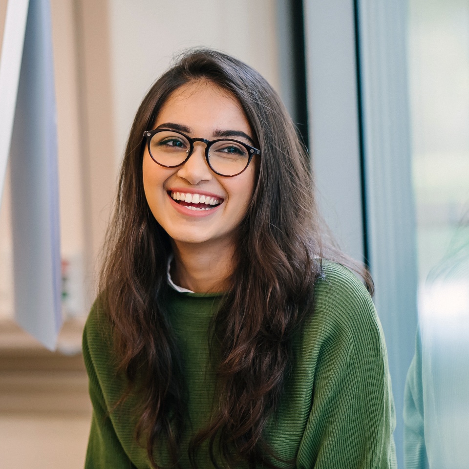 A close up head shot portrait of a preppy, young, beautiful, confident and attractive Indian Asian woman in a green sweater and spectacles in a classroom or office. She is smiling happily.