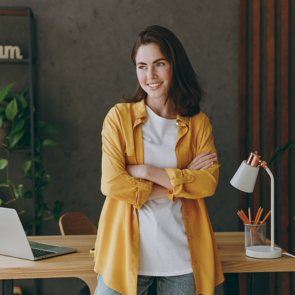 Young minded happy fun calm happy dreamful successful employee business woman 20s she wear casual yellow shirt stand look aside work at wooden office desk with pc laptop. Achievement career concept