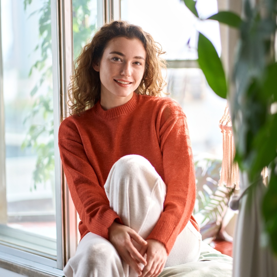 Pretty serene young woman sitting on windowsill relaxing at home looking at camera. Smiling calm lady chilling in apartment, dreaming, thinking of peaceful time enjoying peace of mind. Portrait