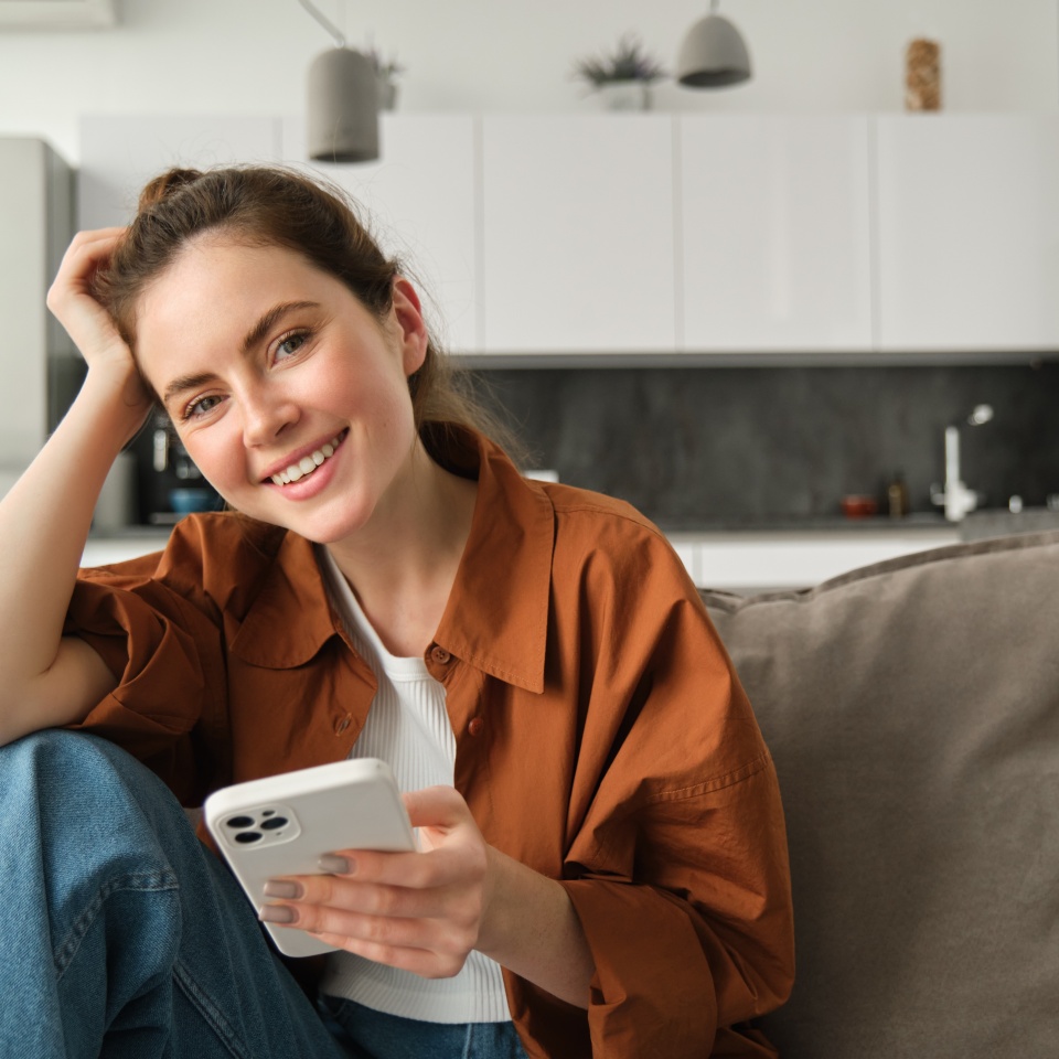 Portrait of cute young smiling woman, sitting on sofa with mobile phone, relaxing at home and using smartphone, scrolling social media, buying online, ordering something on application.