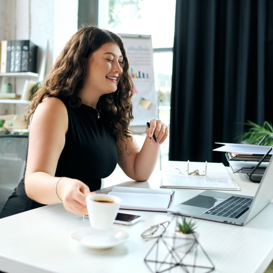 A young plus-size woman with curly hair smiles as she sips coffee at her desk, immersed in her work in a bright office.