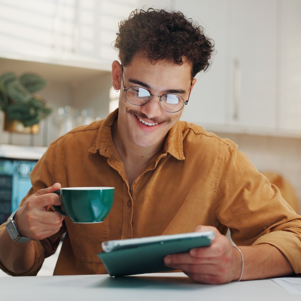 Home, coffee and man with tablet, smile and online reading with morning news, email and internet. Happy, tech and person in kitchen, connection and herbal tea with social media, digital app and latte