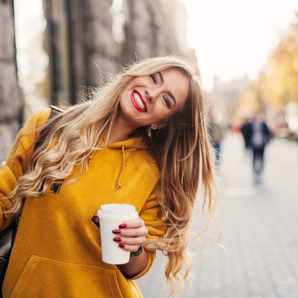 Stylish happy young woman wearing boyfriend jeans, white sneakers bright yellow sweatshirt.She holds coffee to go. portrait of smiling girl in sunglasses and bag