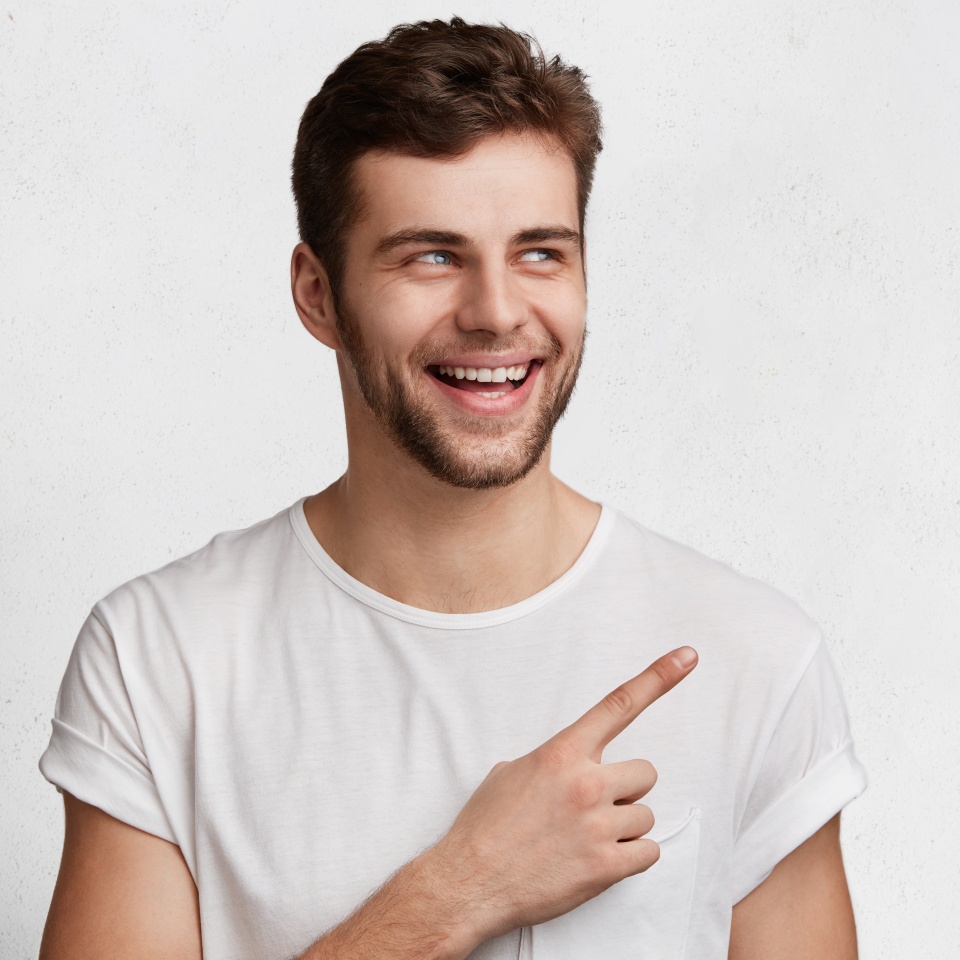 Confident positive male model has appealing smile, looks with joyful expression, isolated over white concrete wall with copy space for advertisment or promotional text. Cheerful man poses in studio