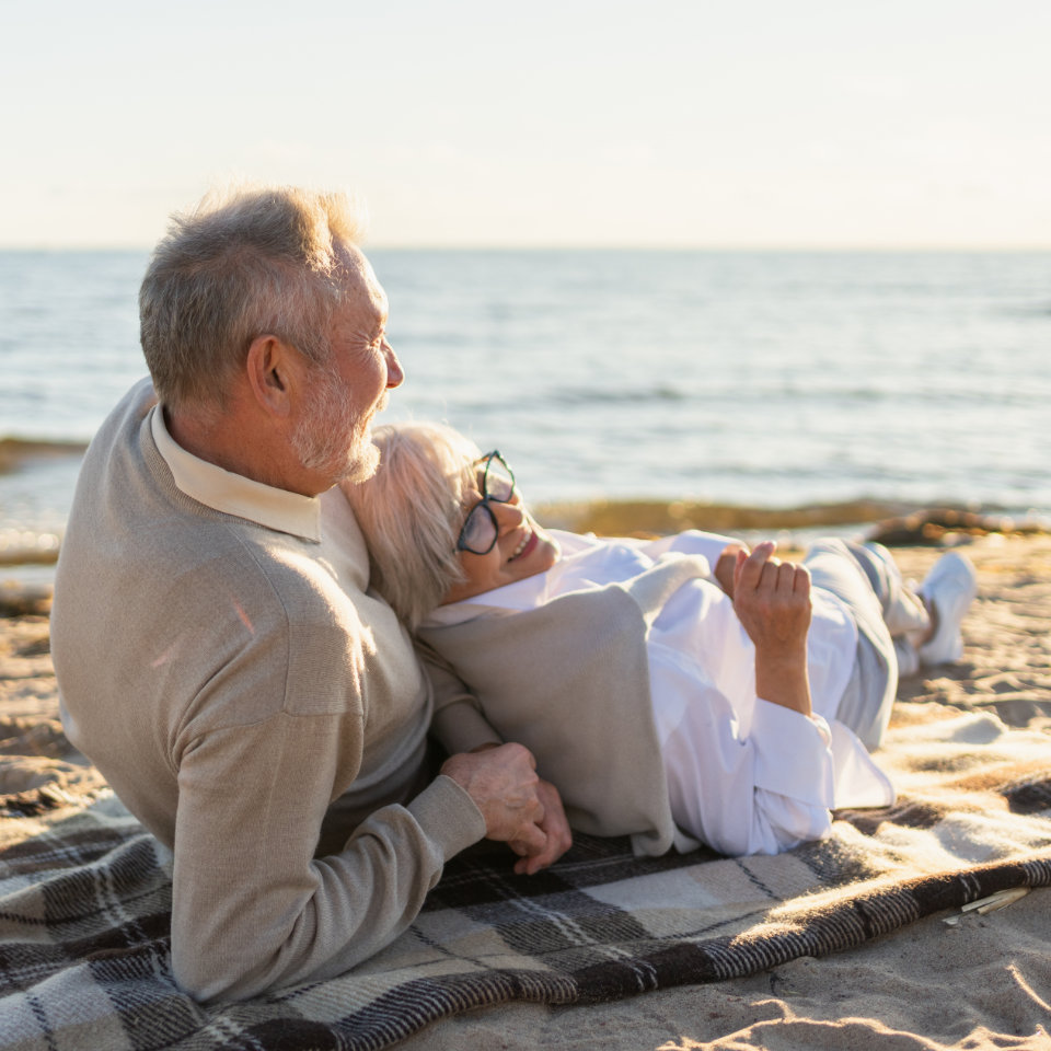 An older couple lies on a blanket at the beach, holding hands and looking toward the water on a sunny day.