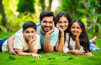 Joyful Indian couple with their children embracing each other and smiling at the camera while sitting in a park, showcasing love, togetherness, and quality family time in a serene outdoor environment