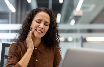 Woman suffering from a painful toothache, holding her jaw and closing her eyes in discomfort, struggling to concentrate on working with her laptop at her office desk